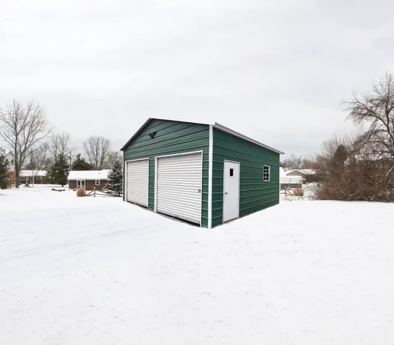 20x20x10 boxed eave roof metal garage with green siding, two roll-up doors, side entry door, and window in a snowy residential setting. This spacious metal garage provides secure storage for vehicles and equipment, and Eagle Carports offers metal garages for sale to protect your property year-round.